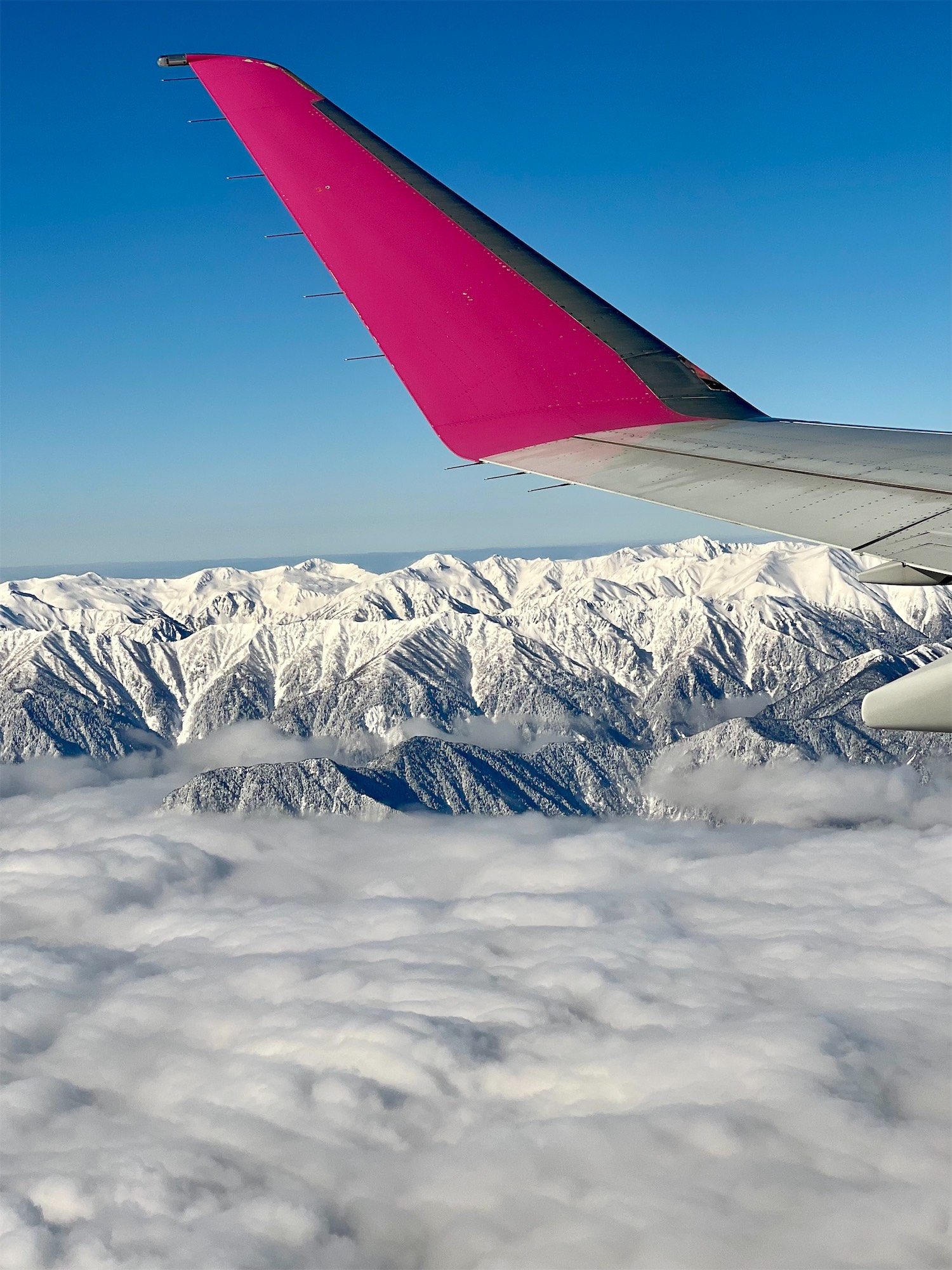 hakuba-plane An airplane wing with a bright pink wing tip flying high above snow-covered mountains and thick white clouds.