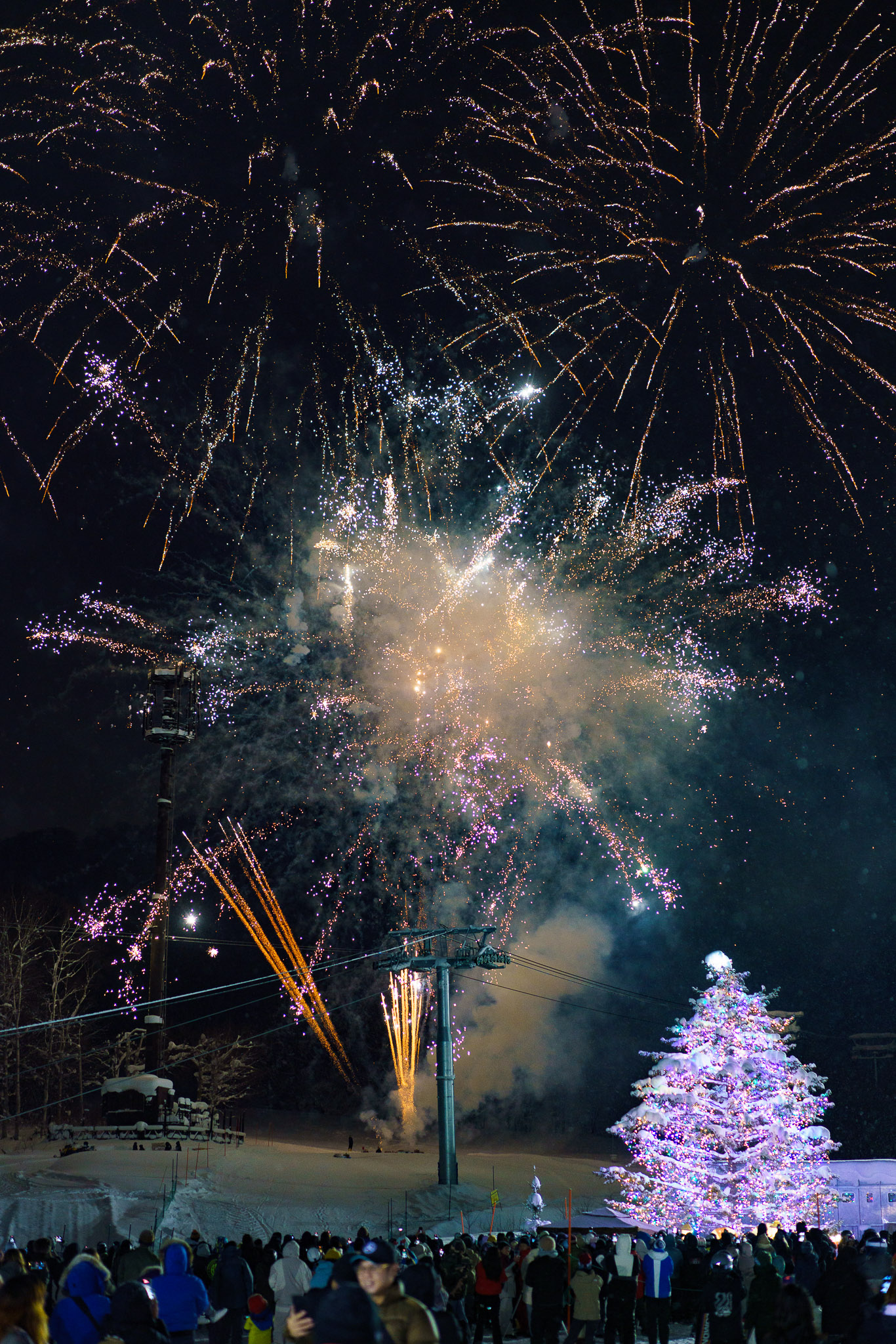 A festive night scene at Niseko Hanazono ski resort, where a crowd of people gathers in the snow. Fireworks explode in the night sky above a brightly lit, decorated Christmas tree. The scene is illuminated by the colorful lights of the tree and the fireworks.
