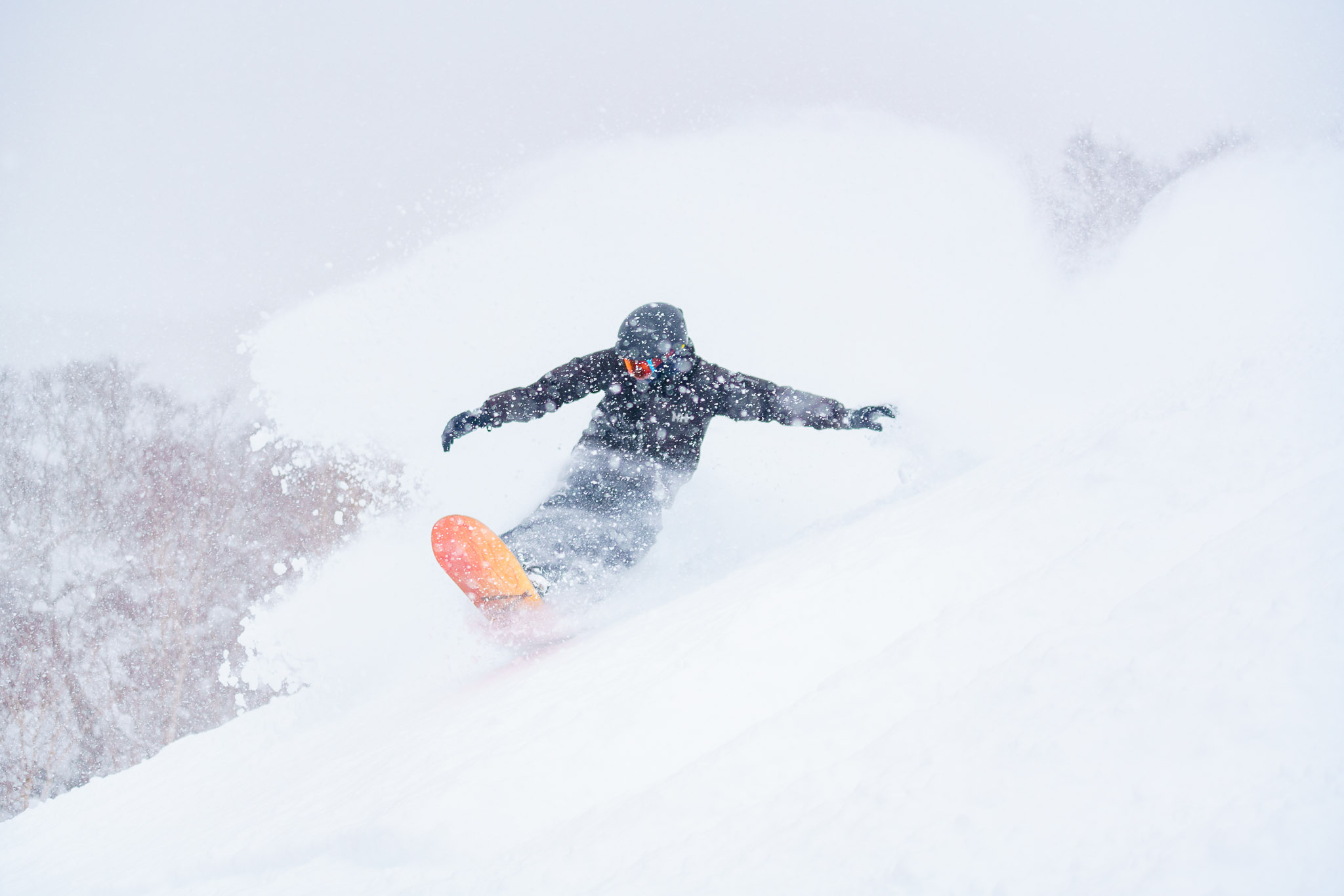 A snowboarder wearing a black jacket and pants is carving through deep powder snow, kicking up a large spray of white snow. The orange bottom of the snowboard is visible as they turn on the snowy slope.