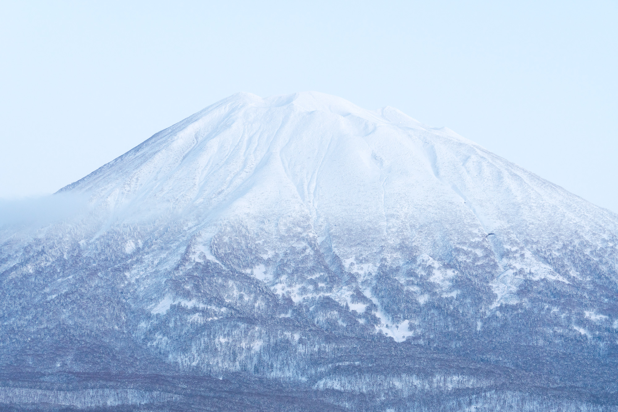 Snow-covered mountain peak with soft blue sky, showcasing winter slopes, frosty ridges, and forested foothills - the backdrop of the new Niseko promo video.