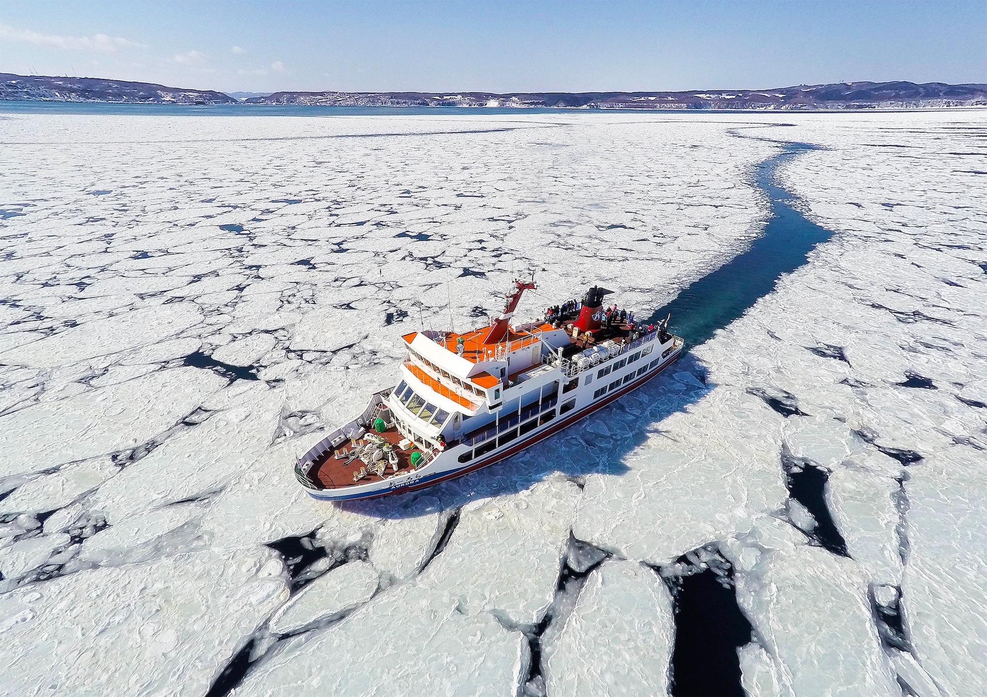 A sightseeing icebreaker ship navigates through thick sea ice under a clear blue sky, carrying passengers across the frozen Sea of Okhotsk during winter in Hokkaido.