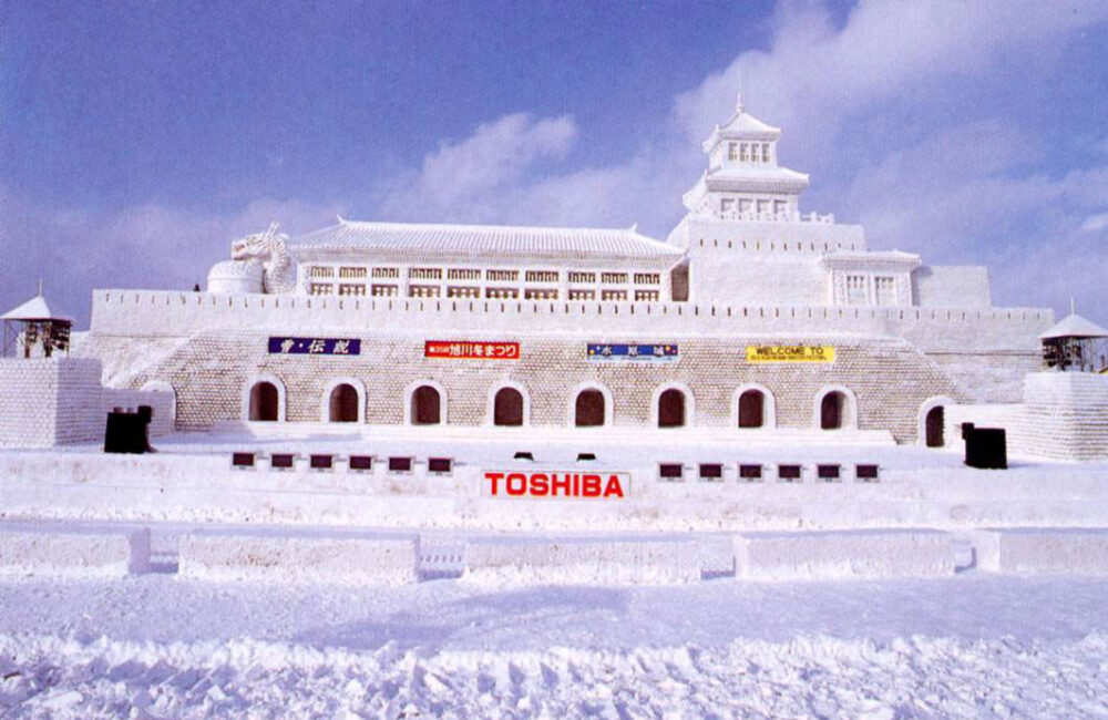A vast snow sculpture shaped like a grand fortress stands under a blue sky at the Asahikawa Winter Festival, showcasing detailed craftsmanship and icy architecture.