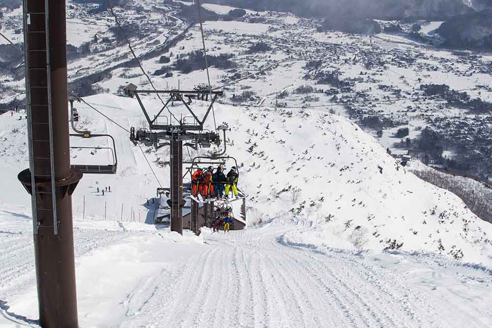 A ski lift carrying skiers and snowboarders ascends a mountain at a resort in Japan, with a snow-covered village visible in the valley below. This image highlights why Hakuba is a premier destination for those wondering where to ski in Hakuba.