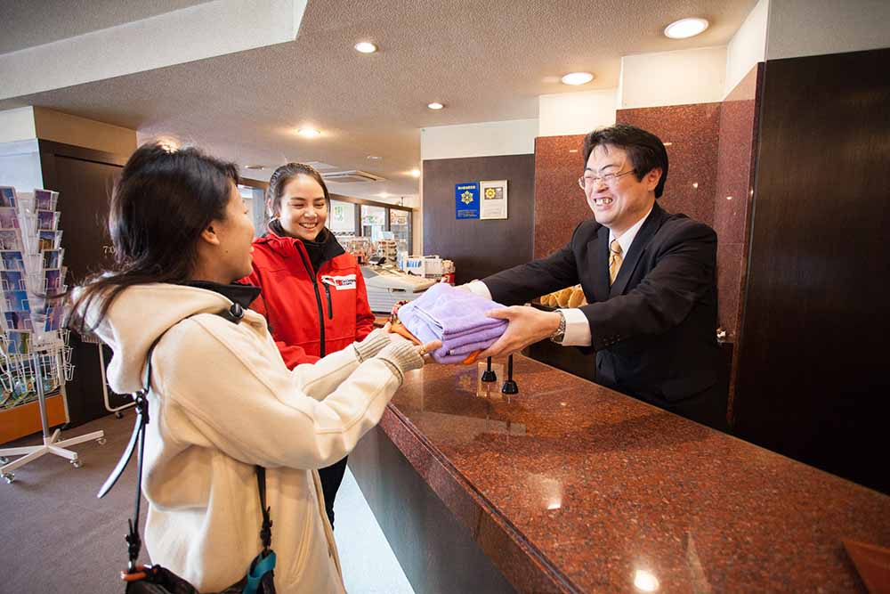 A hotel employee in a suit hands a stack of fresh towels to two smiling guests at a front desk in Japan, showcasing friendly hospitality.