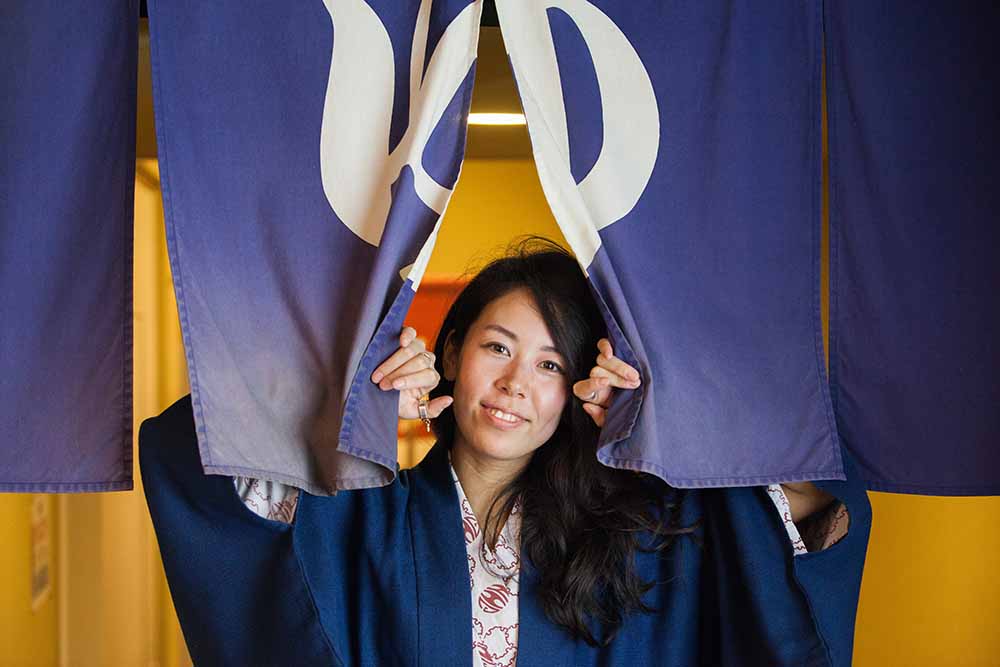 A smiling person in a kimono peeking through a traditional blue noren curtain with the "yu" character, indicating an onsen or hot spring.