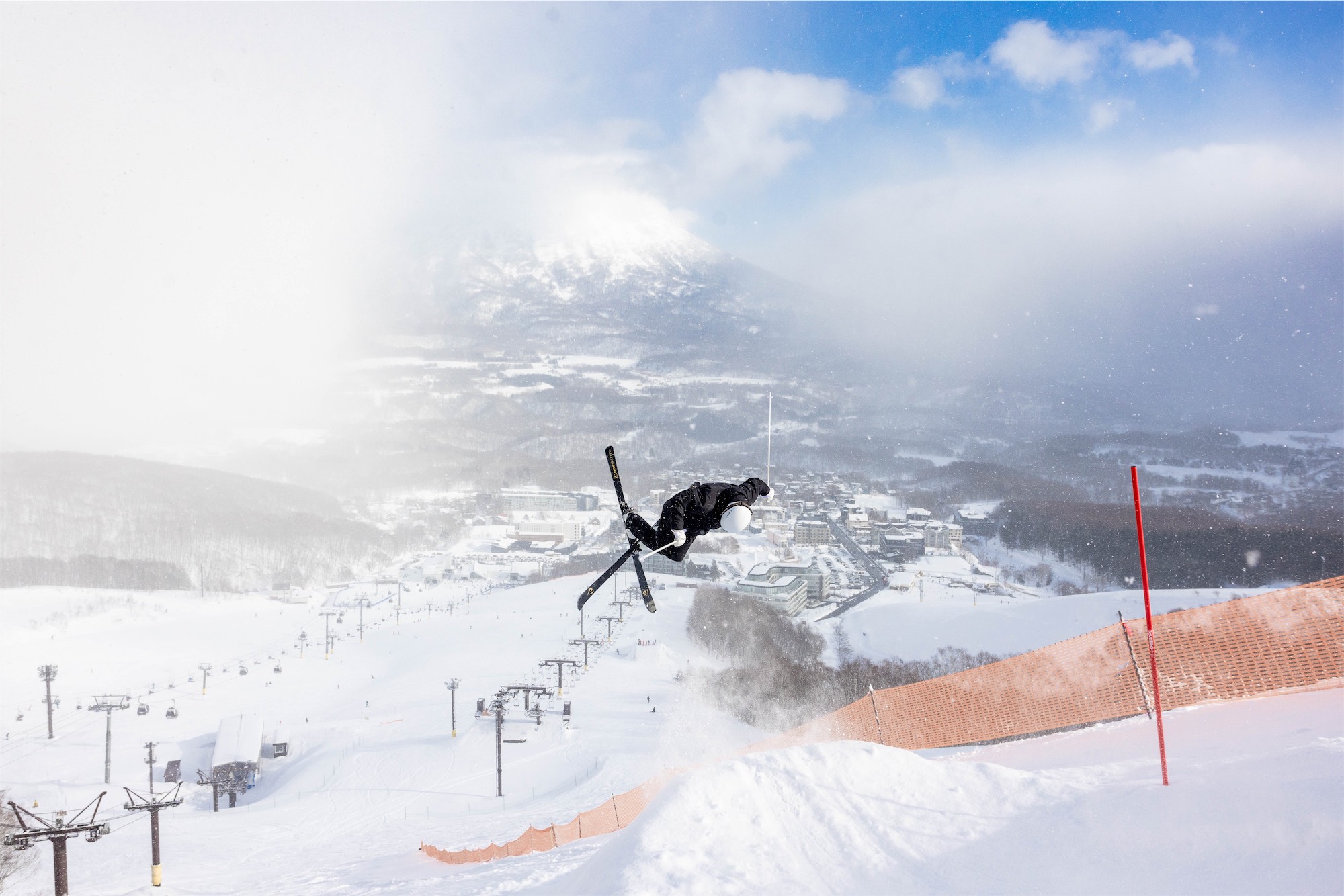 ikuma-horishima-1 Mogul skier Ikuma Horishima doing an aerial trick over a snowy slope with mountain and ski resort in the background.