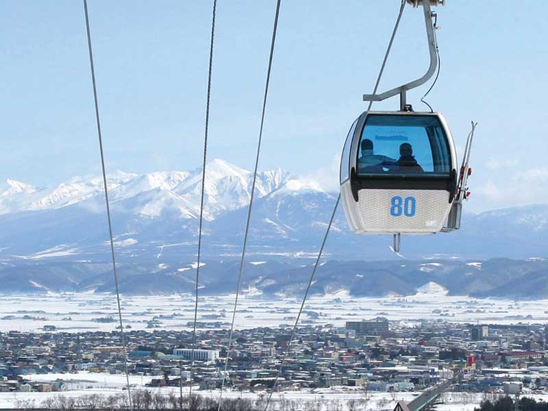 japans-best-ski-resorts-3 Blue cable car ascending above snowy town with distant Japanese Alps in clear winter sky.