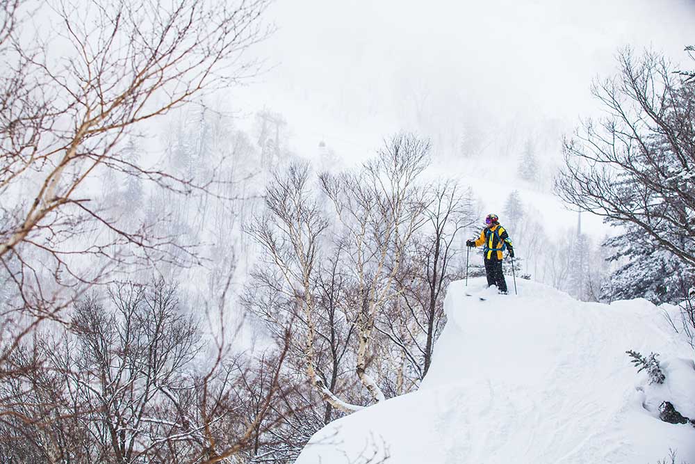 japans-best-ski-resorts-5 Skier standing atop snowy ridge overlooking misty forested mountains in Japan.