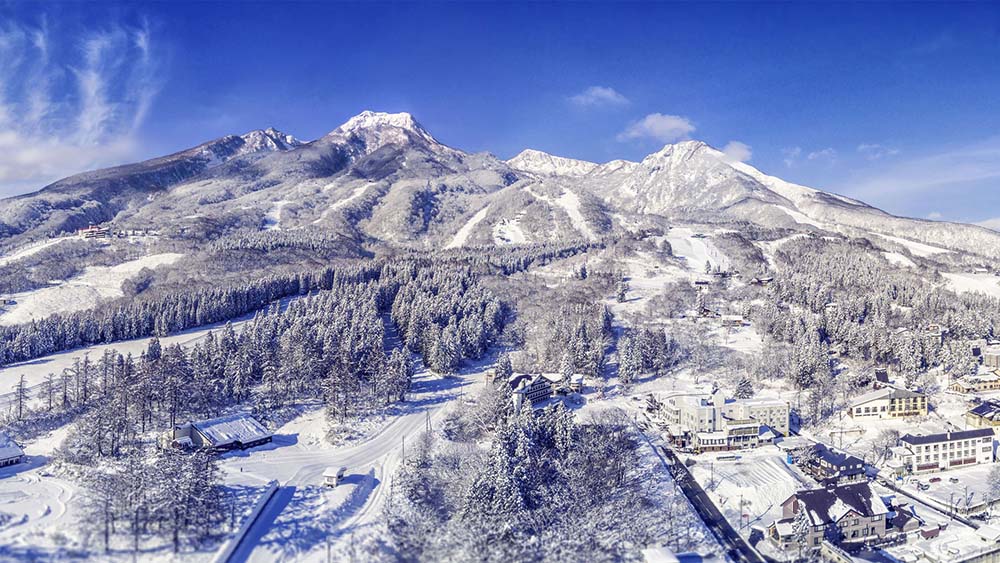 japans-best-ski-resorts-7 Snowy Japanese ski village beneath dramatic mountain peaks on a bright blue winter day.