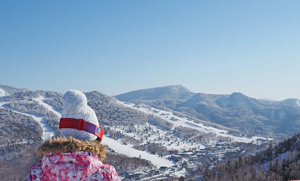 japans-best-ski-resorts-8 Person in winter coat overlooking snowy ski slopes and distant mountains under clear sky.