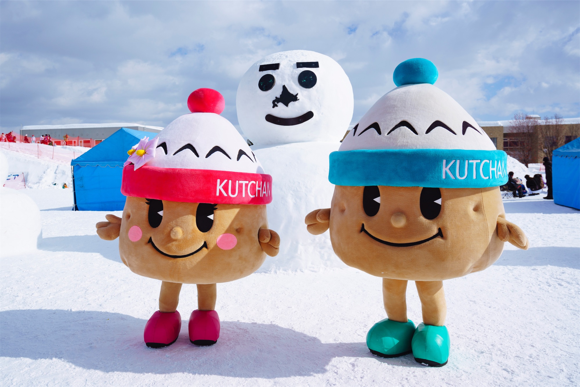 Two cheerful mascots wearing red and blue hats labelled “KUTCHAN” pose in the snow before a large snowman under a bright winter sky at the Kutchan Yukitopia Festival.