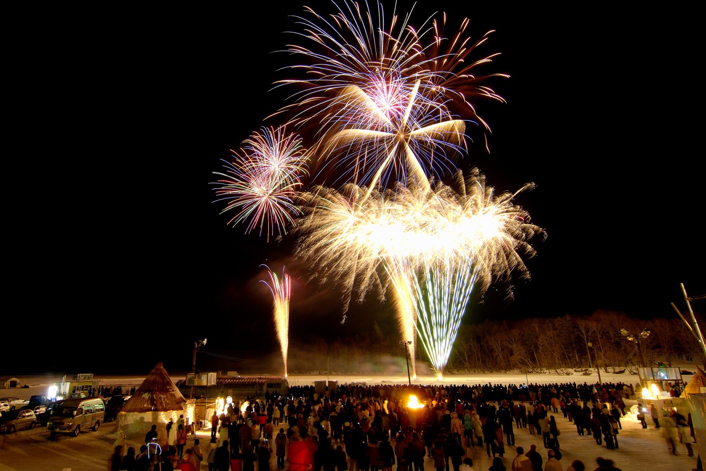 Crowds gather on a snowy field watching colourful fireworks burst across the night sky, celebrating the Lake Akan Ice Festival in Hokkaido, Japan.