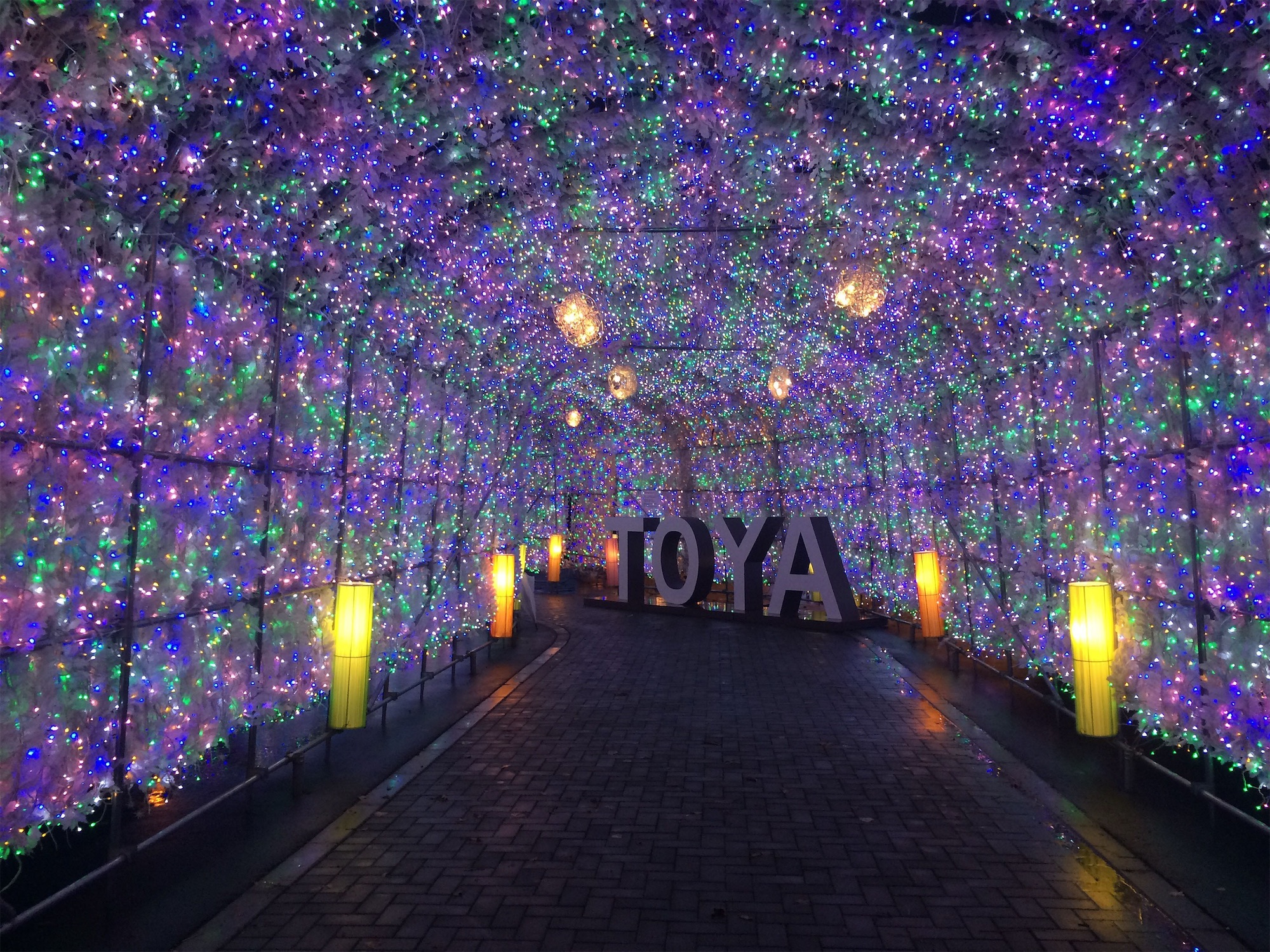 A tunnel of shimmering blue, green, and white lights leads to large “TOYA” letters, creating a dazzling walkway at the Lake Toya Illumination Street in Hokkaido.