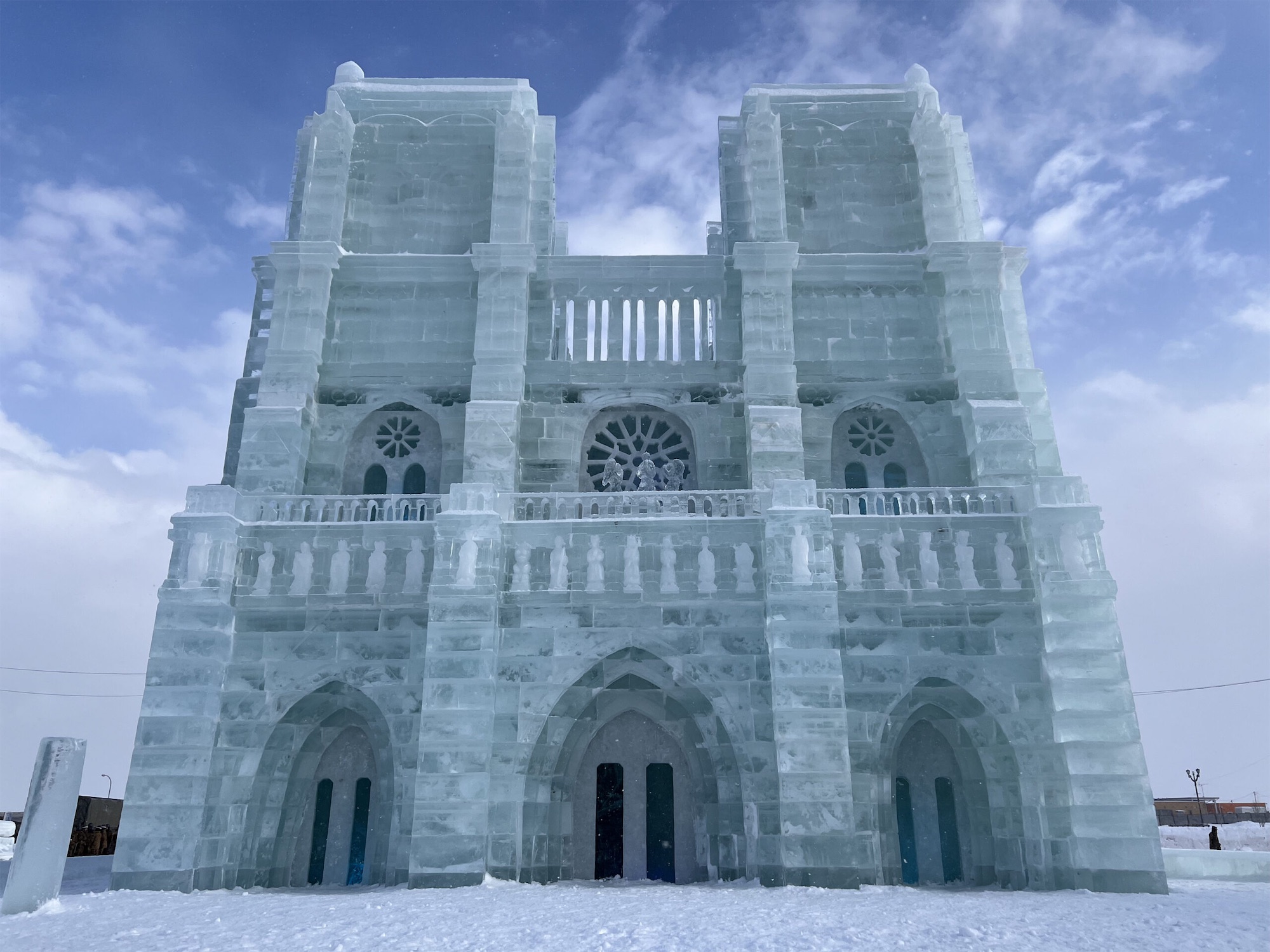 A grand ice sculpture of Notre Dame cathedral stands tall under a partly cloudy sky, showcasing intricate frozen architecture at the Monbetsu Drift Ice Festival.