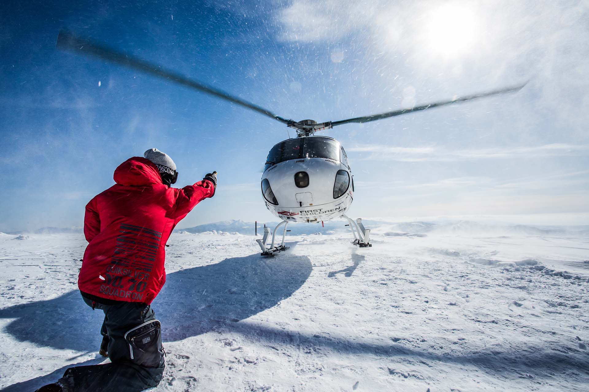 niseko-heli-skiing-1 Guide in red jacket directing helicopter landing on snow-covered ridge under bright blue sky.