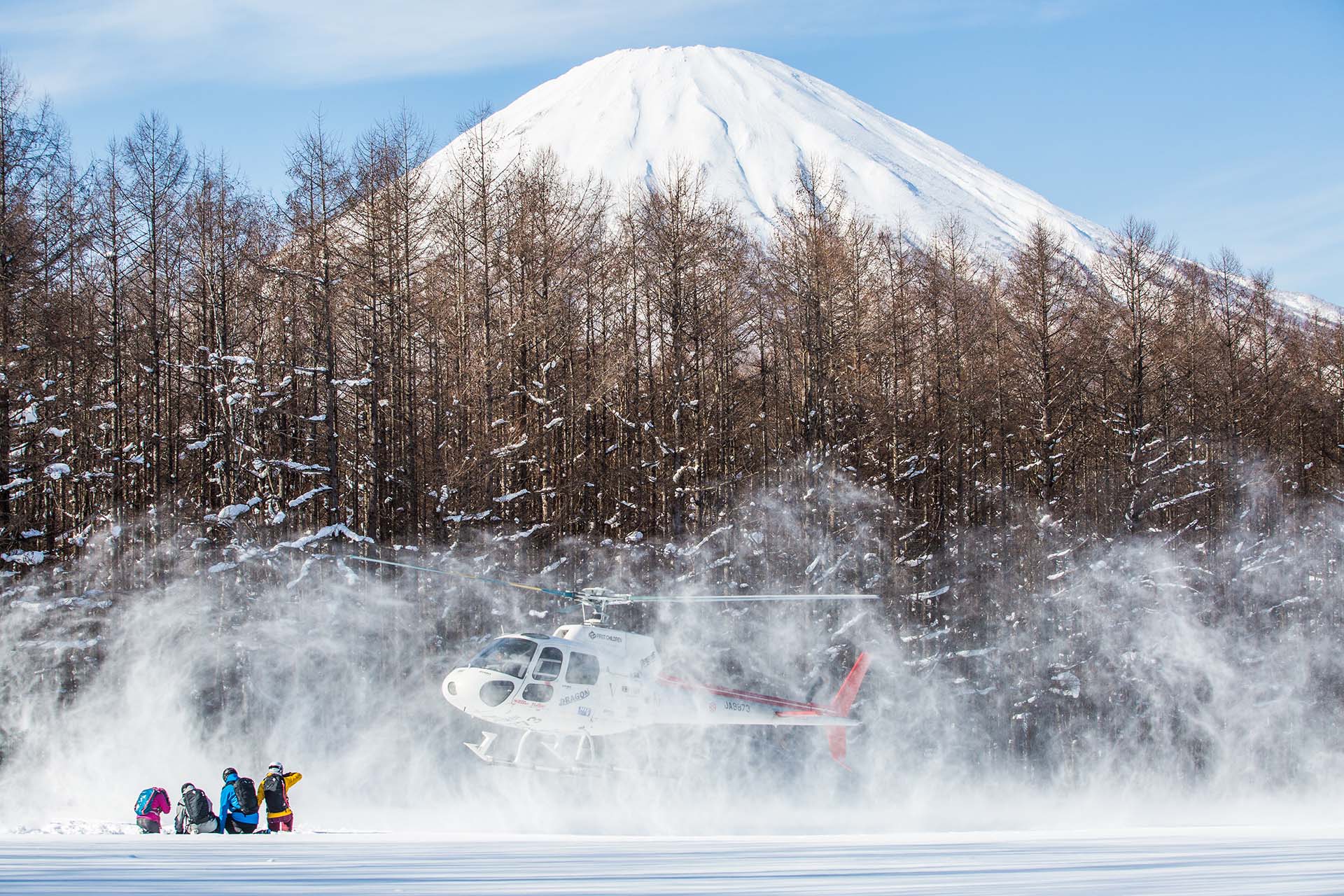 niseko-heli-skiing-3 Helicopter landing in snowy forest clearing near Mount Yotei with skiers preparing for heli-ski adventure.