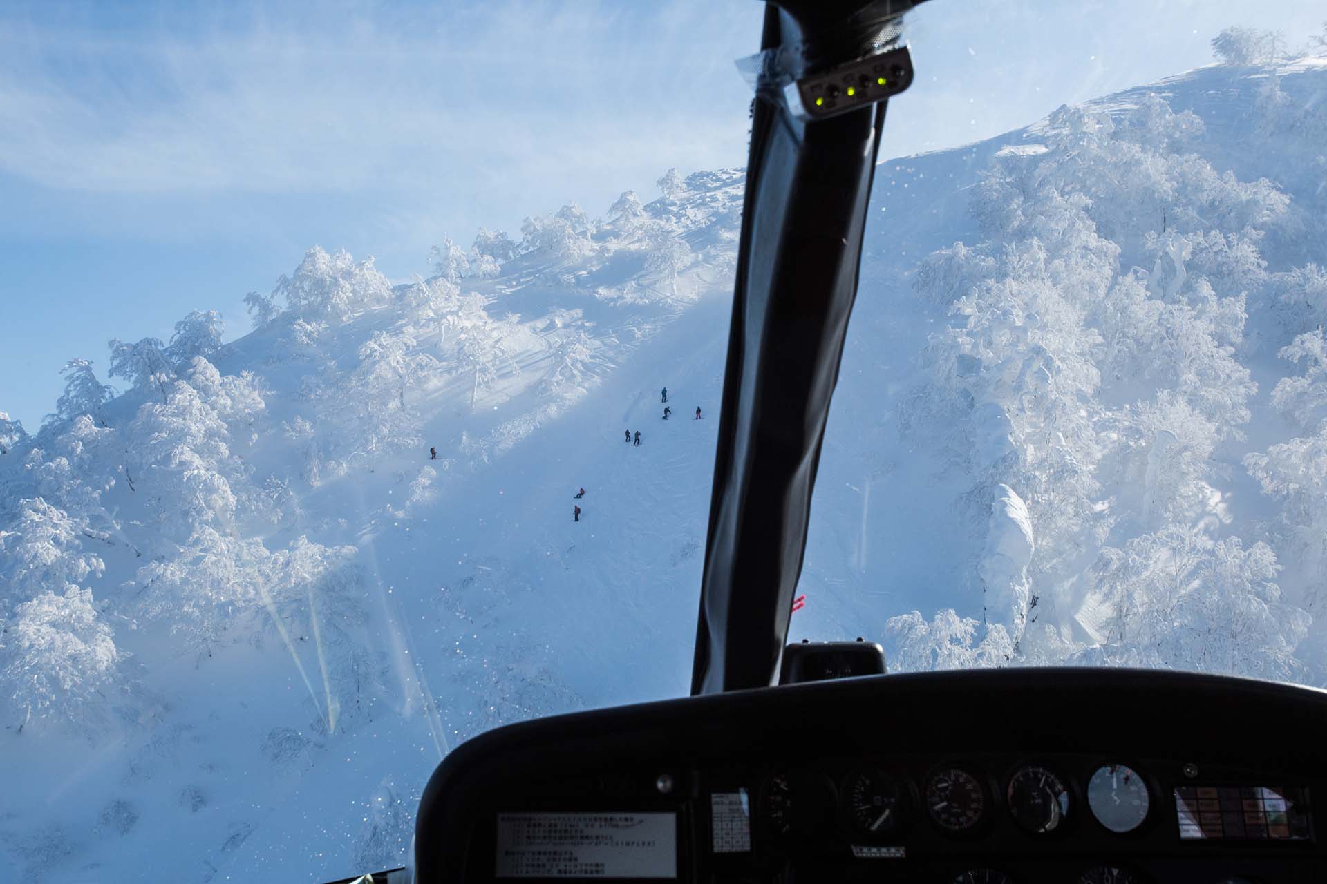 niseko-heli-skiing-4 View from helicopter cockpit flying over snow-covered mountain slope with skiers descending below.