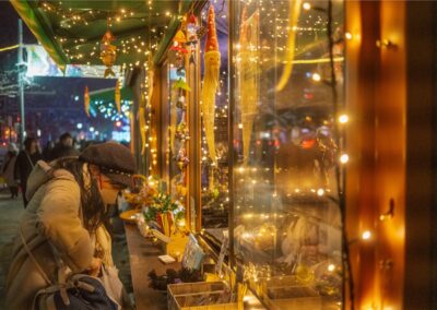 A person wearing a hat and face mask browses a brightly lit Sapporo Christmas Market stall at night, adorned with string lights.
