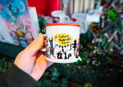 Hand holding a white mug with red rim and colorful 2025 Munich Christmas in Sapporo illustration