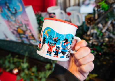 Hand holding a white mug with red rim and colorful 2025 Munich Christmas in Sapporo illustration.