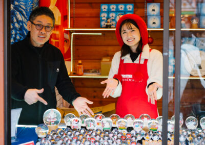 Man and woman in a shop, smiling and presenting an extensive collection of snow globes on display.