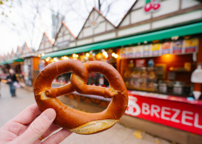 A hand holds a large, golden-brown pretzel in the foreground of an outdoor market stall with a 'BREZEL' sign.