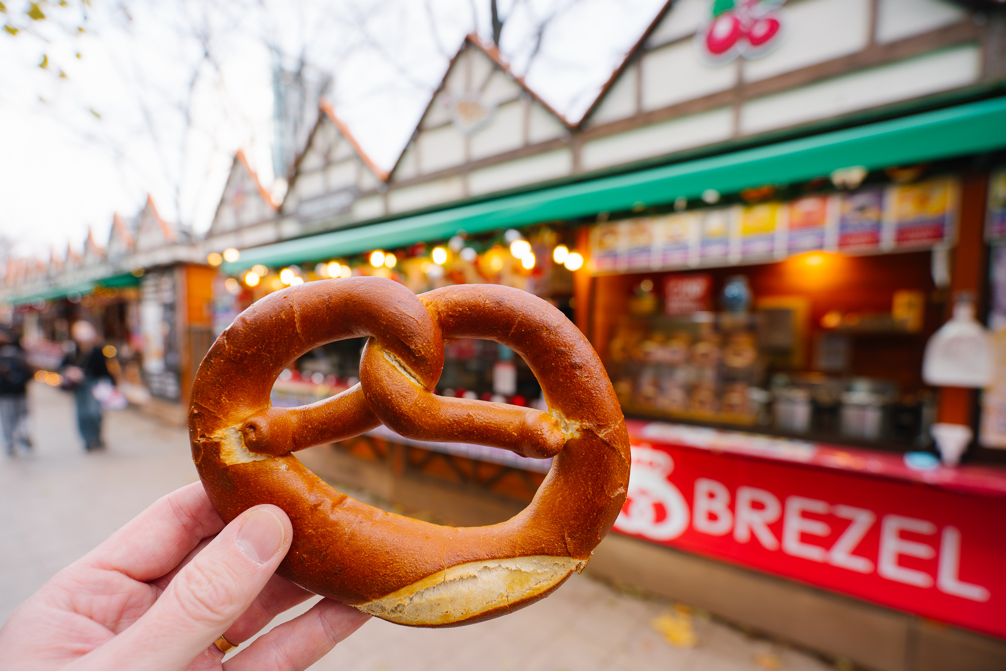 sapporo-christmas-market-2025-4 A hand holds a large, golden-brown pretzel in the foreground of an outdoor market stall with a 'BREZEL' sign.