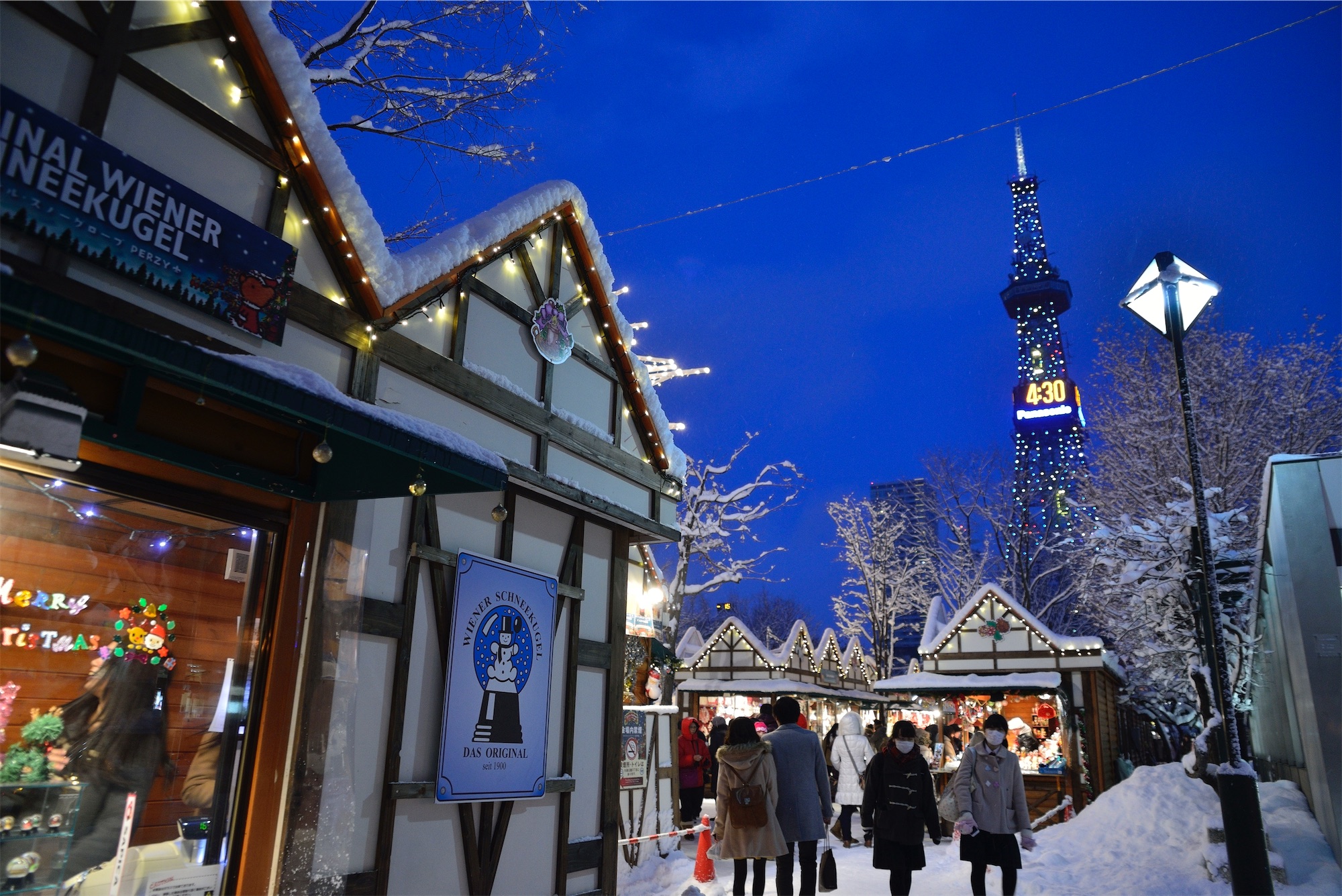 People stroll through a snowy Christmas market at dusk in Sapporo, Japan, with festive lights and stalls glowing beneath the illuminated TV tower.
