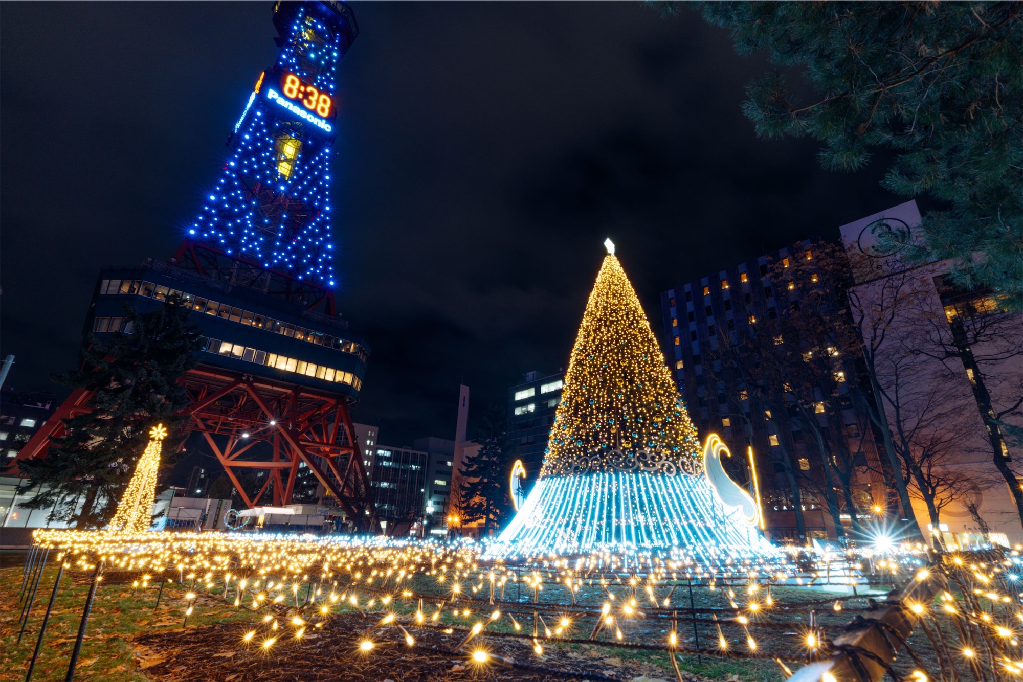 A large illuminated Christmas tree and glowing light displays shine beside Sapporo TV Tower at night, creating a festive winter scene at one of the top Hokkaido snow festivals.