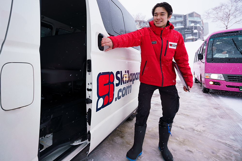 Skiers ride a snowy mountain chairlift above wide powder fields with distant hills and winter landscape in soft, cloudy light.