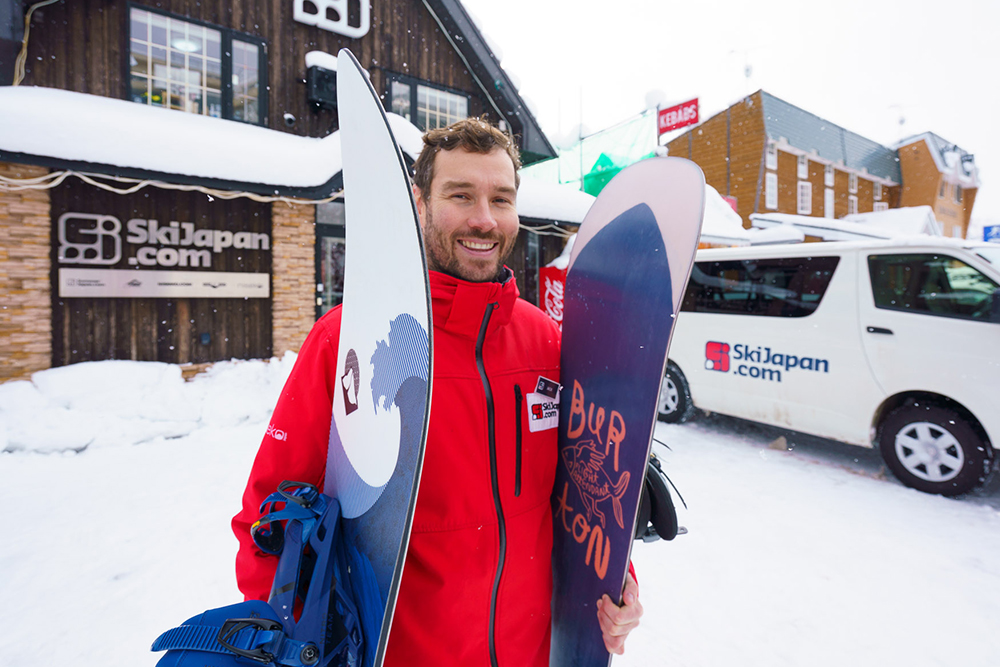 Skiers ride a snowy mountain chairlift above wide powder fields with distant hills and winter landscape in soft, cloudy light.
