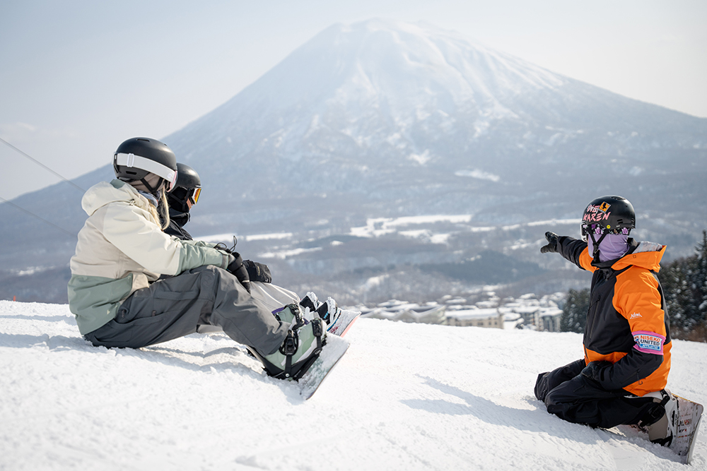 Skiers ride a snowy mountain chairlift above wide powder fields with distant hills and winter landscape in soft, cloudy light.