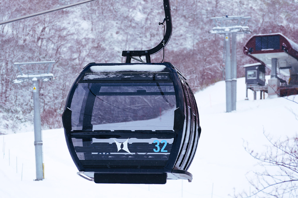 Skiers ride a snowy mountain chairlift above wide powder fields with distant hills and winter landscape in soft, cloudy light.